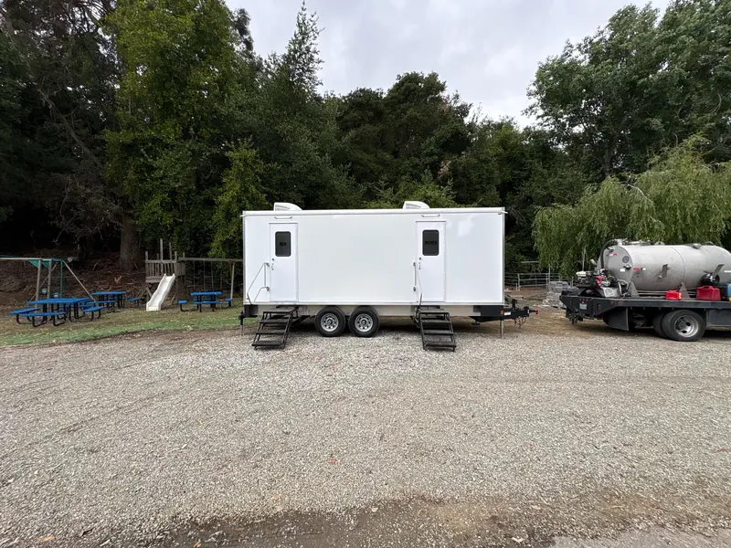 Restroom trailer on grass under shade trees at a park event